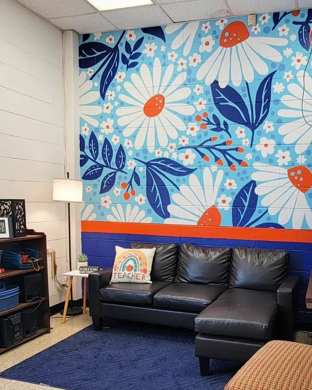 Colorful mural of large white daisies with orange centers, blue leaves, and small white and orange flowers on a blue background in a classroom corner with a black leather couch, a pillow that says 'TEACHER', a small white side table, and a bookshelf.