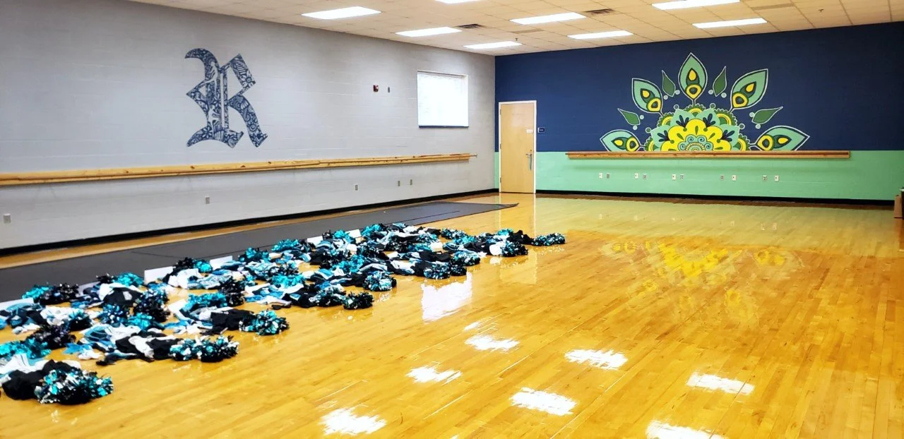 Indoor dance studio with a large, colorful floral mural on the right wall and a blue letter 'R' on the left wall. Cheerleading pom-poms are scattered on the shiny wooden floor in the foreground.