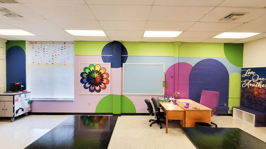 Colorful classroom with mural wall art, featuring geometric shapes, a framed colorful flower design, a whiteboard, desk with office supplies, and a purple armchair with a decorative poster reading 'Love One Another' on the wall.