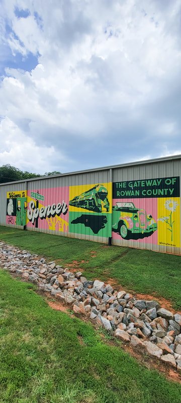 Colorful mural on a building wall welcoming visitors to Rowan County, featuring a train, a vintage truck, and floral designs under a cloudy sky.