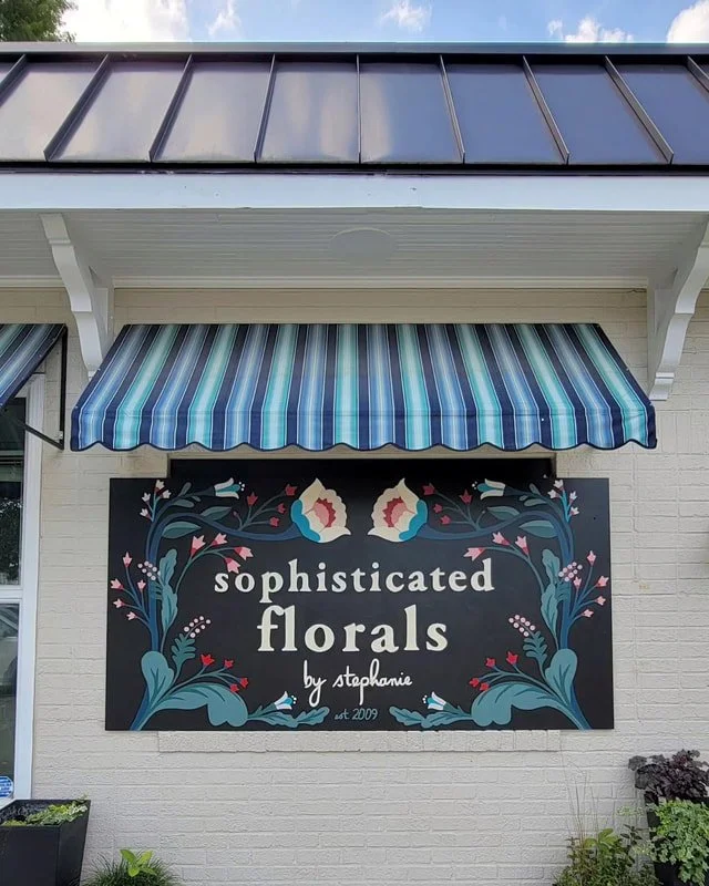 Sign for a floral shop called 'sophisticated florals by stephanie' with decorative floral artwork, installed on a white brick wall below a striped blue and gray awning, part of a building with a metal roof.