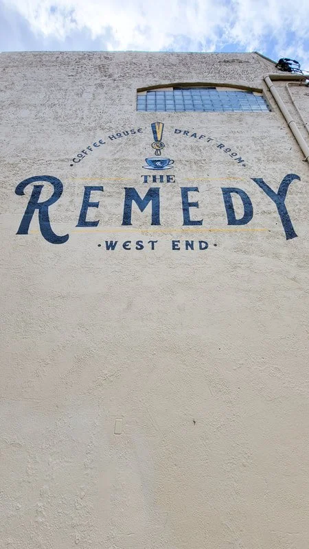 Sign on a building for The Remedy, a coffee house and draft room in West End, featuring a logo with a cup of coffee and a spoon.