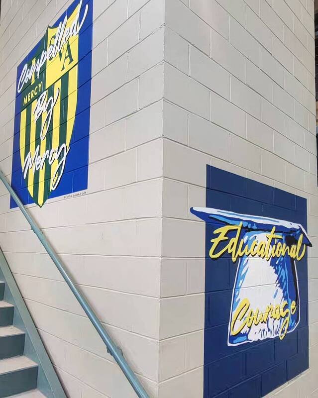 Decorative school-themed murals on a beige brick wall, featuring a shield with yellow and green stripes and the words 'Courage, Mercy, Mercy' and a graduation cap with the words 'Educational Courage'.
