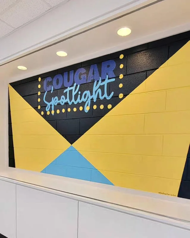 A painted wall with the words "COUGAR spotlight" in large letters, with "COUGAR" in blue and "spotlight" in light blue cursive, surrounded by yellow and black triangular shapes and yellow dots.