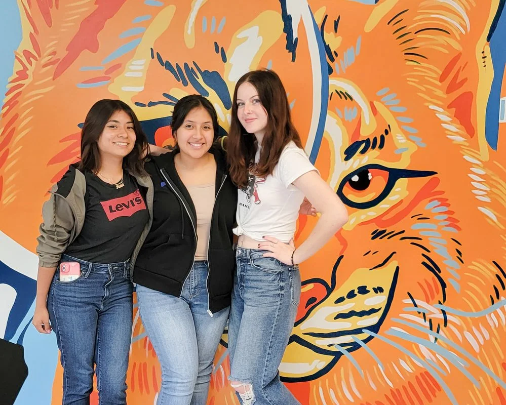 Three young women standing together in front of a vibrant orange tiger mural, smiling and posing for the photo.