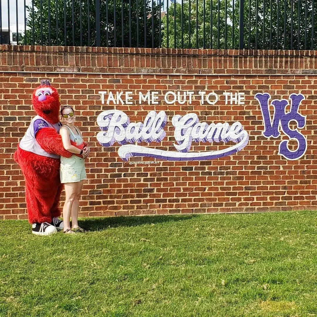 A person standing next to a mascot character dressed as a furry red creature with a purple nose, on a grassy area in front of a brick wall with painted purple and white text that reads, "Take me out to the Ball Game VS".
