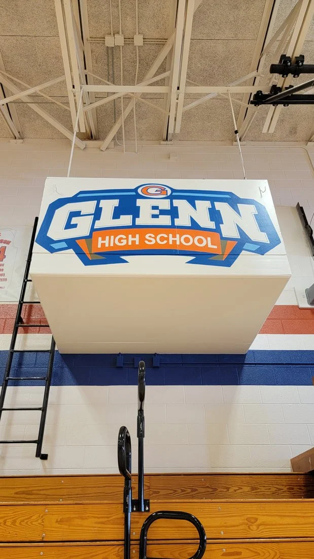 A basketball gymnasium ceiling featuring a large egg-shaped sign with the logo and name of Glenn High School, adorned with blue, white, and orange colors, and a basketball hoop with a backboard visible at the bottom of the image.