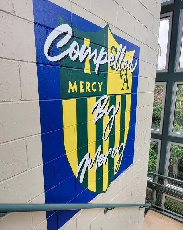 Colorful wall mural featuring the Mercy High School logo with the words "Compliment A Mercy" written in white cursive over a shield-shaped emblem with yellow and green vertical stripes.
