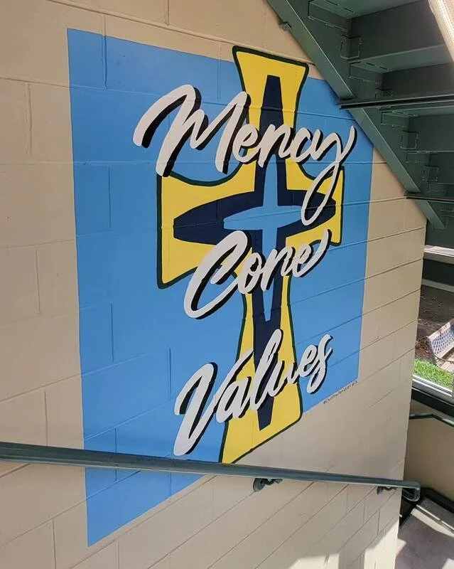 A mural with the message "Money  Gone  Values" on a wall near a staircase, featuring a large yellow cross with a blue and black design in the background.