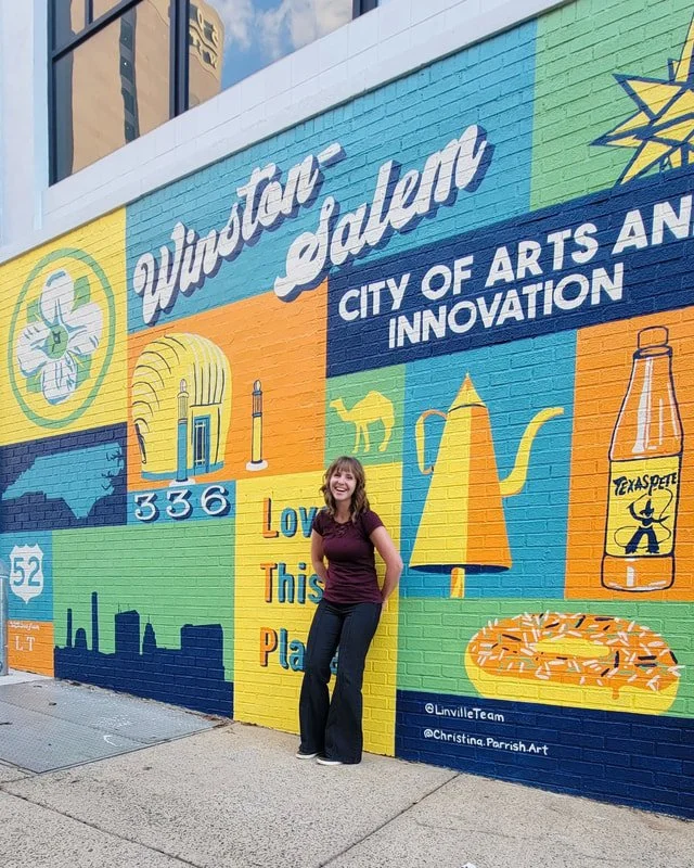 A woman standing in front of a colorful mural on a brick wall in Waxahachie, Texas. The mural features images of a flower, a tower, a silhouette of a moon and star, a lamp, a bottle with a logo, a city skyline, and the text 'Waxahachie, Texas' and 'City of Arts and Innovation.
