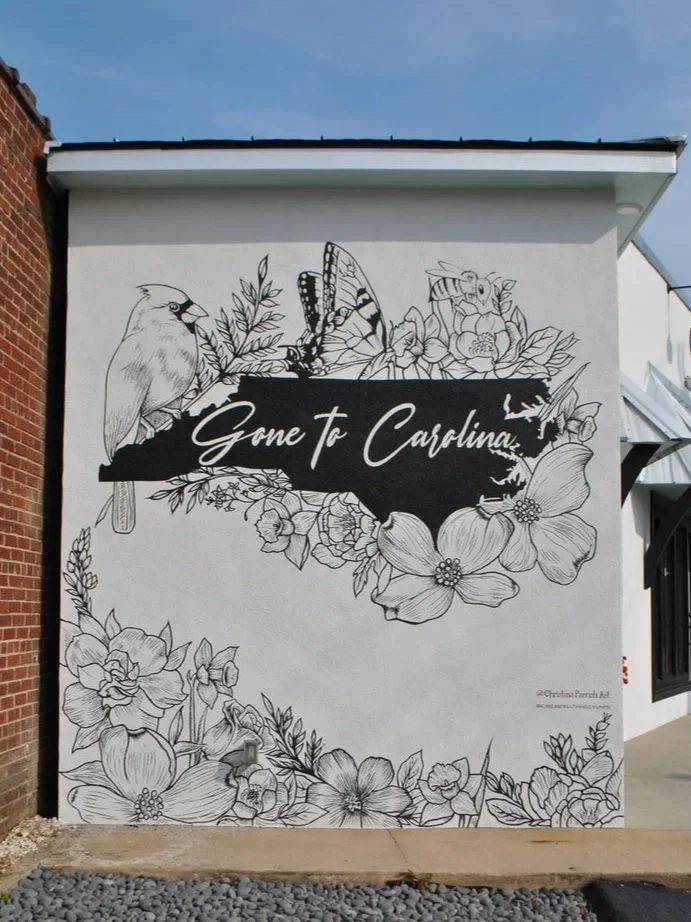 Black and white mural with floral designs, a bird, a butterfly, and a bee surrounding the words 'Gone to Carolina' on a wall.