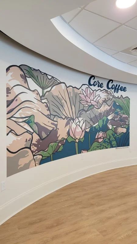 Colorful mural on wall featuring mountains, flowers, and water lilies with the words "Core Coffee" above it, in an indoor space with wooden flooring.