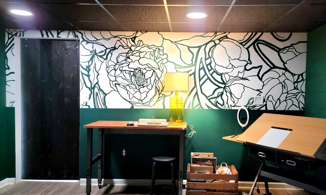 Room with a black door, a wall decorated with a large black and white floral mural, a wooden table with a yellow lamp, a ring light, a small stool, and a wooden tray holding art supplies.