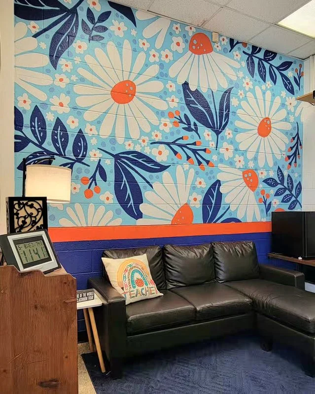 A wall mural with large white daisy flowers, blue leaves, and small orange accents on a light blue background in a cozy waiting area. There is a black leather sofa, a white pillow with a rainbow design, a small side table with a book, a digital clock