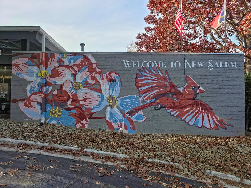 A mural on a brick wall in New Salem features colorful flowers and a cardinal bird, with American flags above.