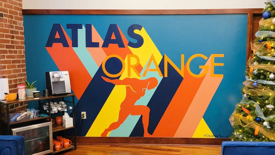 Colorful mural with the words 'Atlas Orange' and an orange silhouette of a person lifting a large sphere, with a Christmas tree decorated with lights and ornaments to the right.