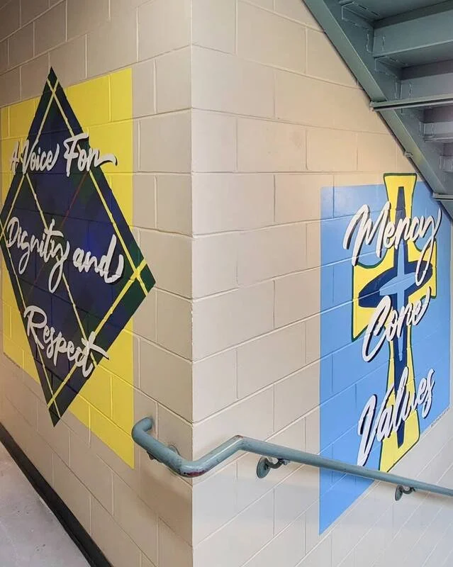 Colorful school hallway with murals. One mural displays the words 'Voice for Dignity and Respect' on a yellow background with a diamond shape. Another mural shows 'Menu and Values' on a blue background with a sword emblem. Handrail runs along the sta