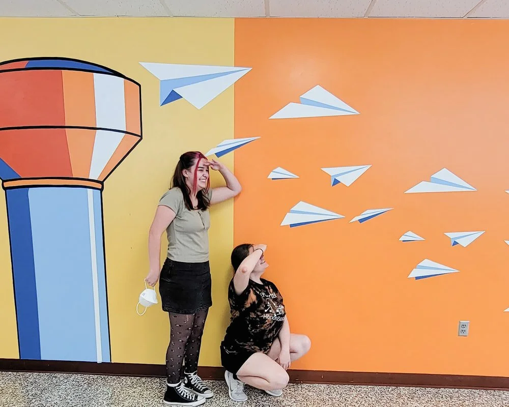 Two women interact with a colorful wall mural that features a large soda dispenser and numerous paper airplanes flying out of it.