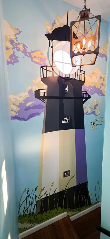 Painted mural of a lighthouse on a wall with a decorative chandelier above. The lighthouse has black, white, and purple sections, a small balcony near the top, and a window. The painting includes clouds, a lighthouse light, and a seagull in the sky.