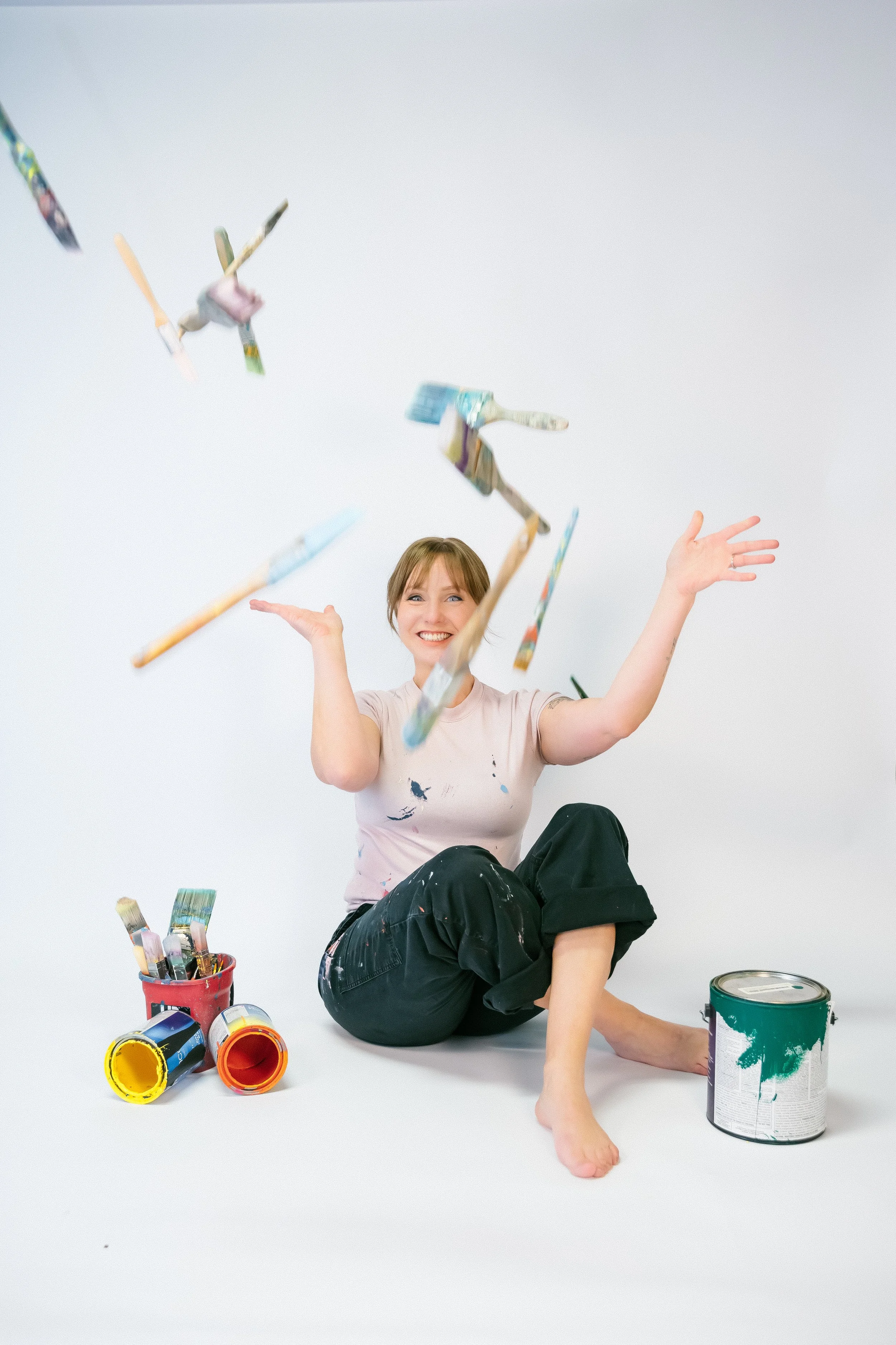 A smiling woman sitting on the floor with her legs crossed, surrounded by open paint cans, a paintbrush holder, and paintbrushes. She is throwing paintbrushes into the air and is wearing a paint-splattered shirt and black pants.