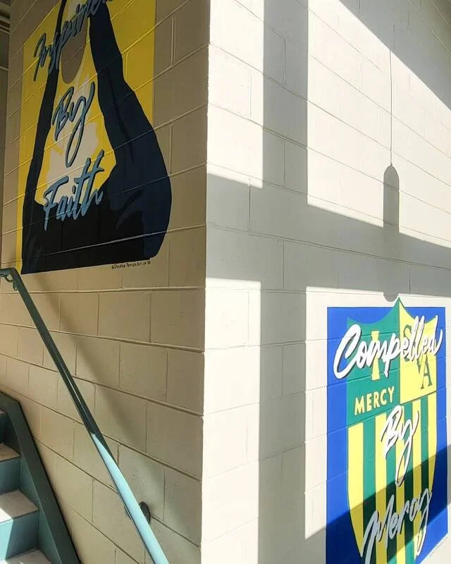 Campbell's Mercy Bull of Mercy sign on a light-colored cinder block wall at an angle with stairs and a railing nearby.