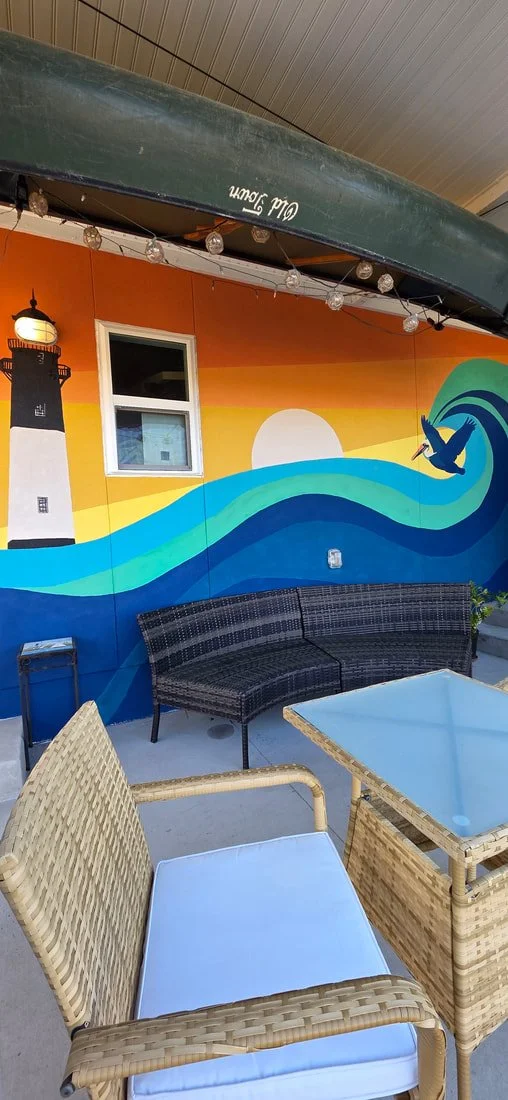 Colorful outdoor patio with a wall mural depicting a lighthouse, a sunset, waves, and a flying bird. There are wicker chairs with cushions, a glass-topped table, and a canoe hanging from the ceiling.