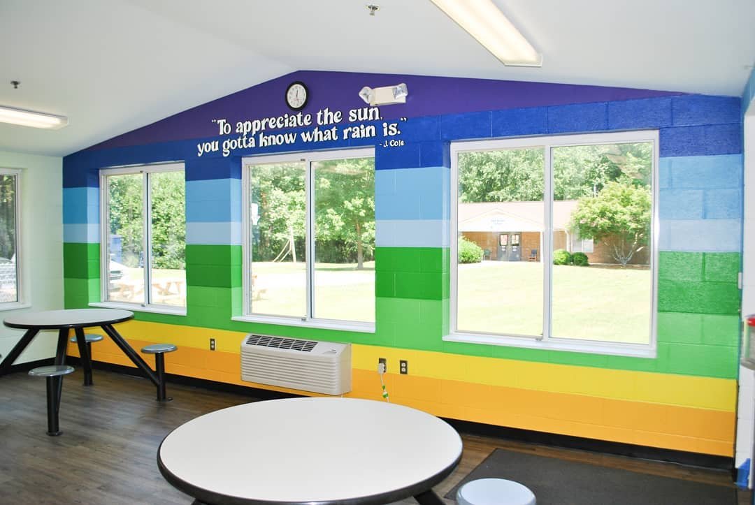 Colorful mural with quote on a school or community room wall, featuring blue, green, yellow, and gray stripes, with three windows showing green trees outside, a clock, and a wall-mounted air conditioner nearby.