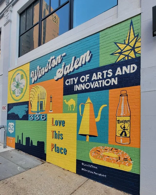 Colorful mural on a city building wall celebrating Winston Salem with symbols of arts and innovation, including a star, cactus, teapot, doughnut, and a bottle labeled Texas Pete, with the words "Love This Place" and city skyline silhouettes.