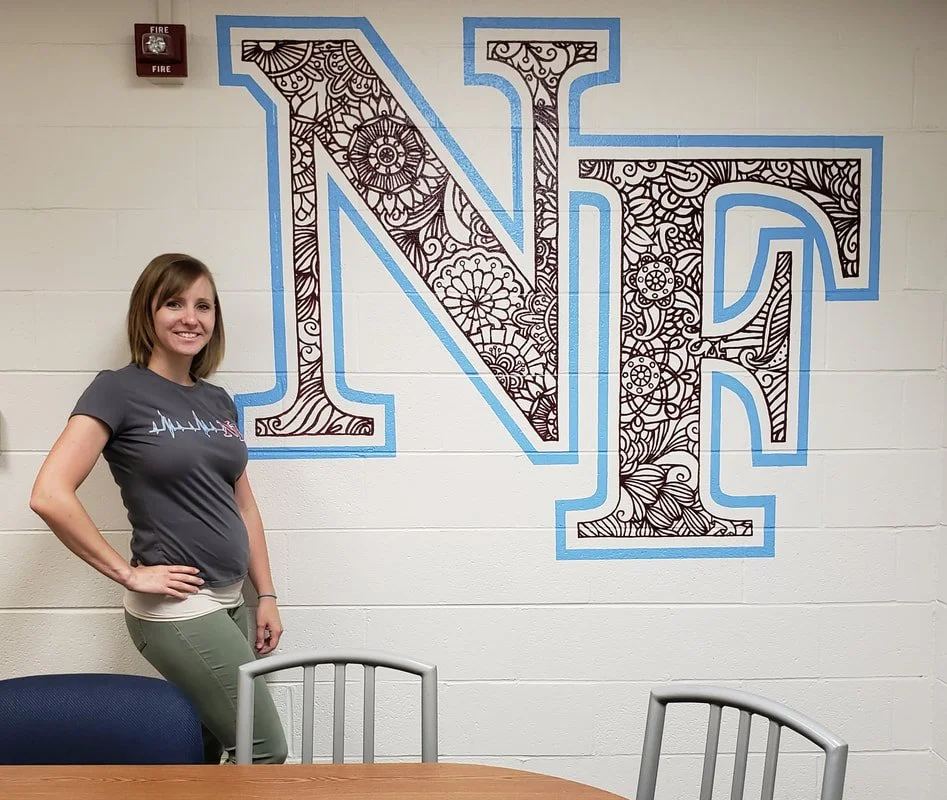 A woman with short hair wearing a gray shirt and light-colored pants standing next to a table with chairs, in front of a large decorated letter N and E on a white wall.