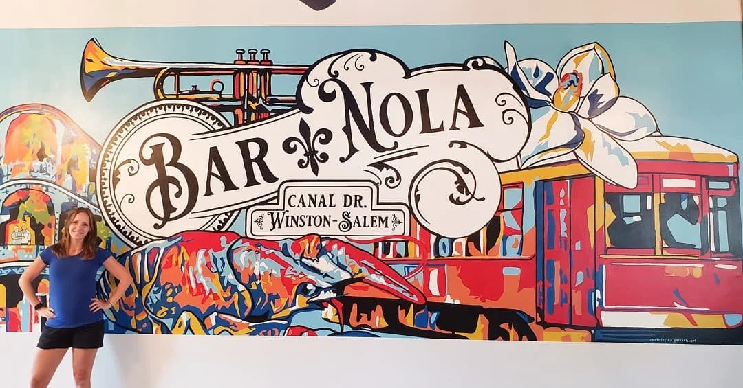 A woman with brown hair, wearing a blue t-shirt and black shorts, stands with her hands on her hips in front of a colorful mural. The mural features the text 'BAR NOLA,' a trumpet, a flower, a streetcar, and the address 'Canal Dr. Winston-Salem.'
