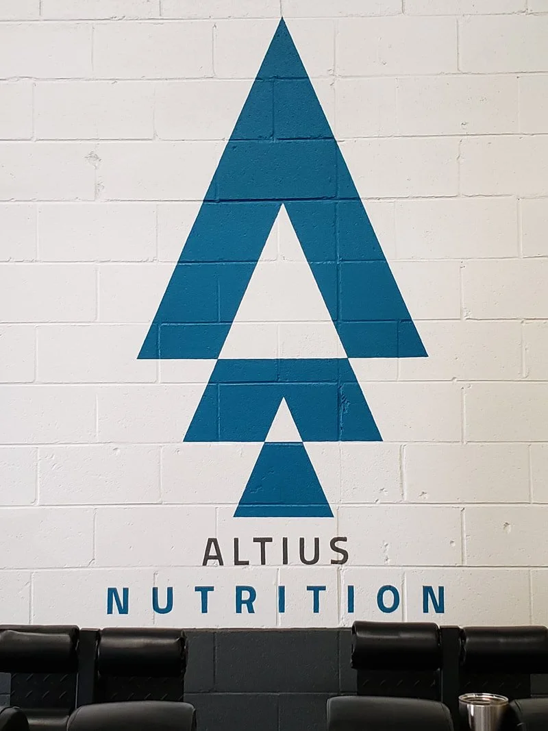A white brick wall features a large blue stylized tree logo with the words "Altius Nutrition" underneath. Black chairs are visible at the bottom of the image.