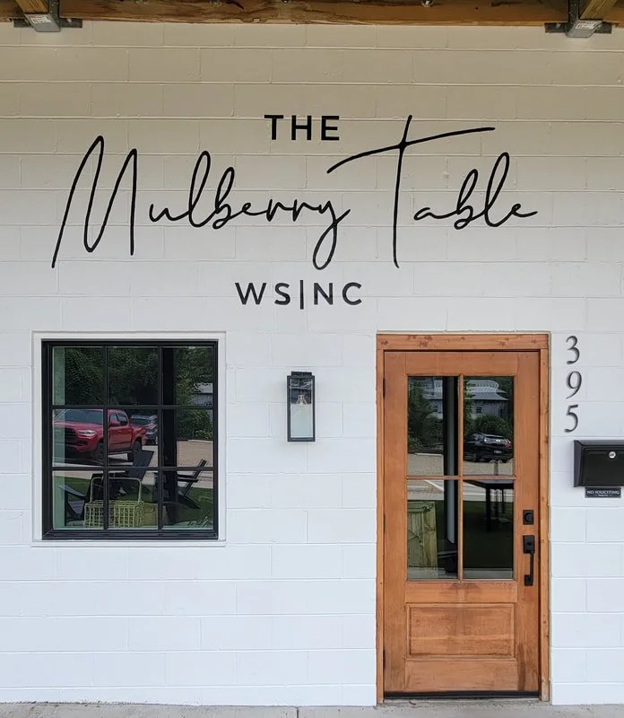 Exterior of a building with a white brick facade and a wooden front door, labeled 'The Mulberry Table WINSC'. The building has a window showing reflections of parked cars and outdoor furniture, and the street address '395' displayed vertically beside