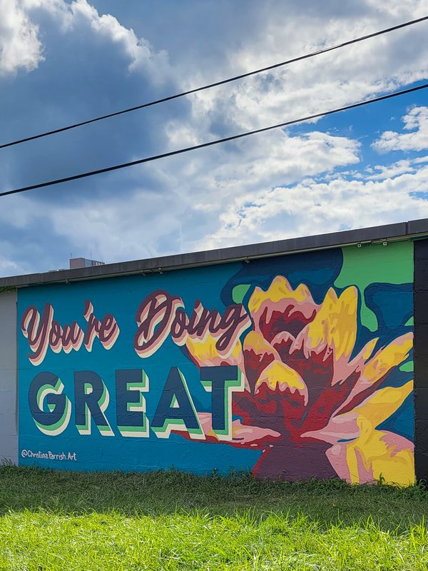 A colorful mural with the words "You're Doing Great" on a blue background, next to a large abstract flower painting, located on the side of a building with power lines overhead.