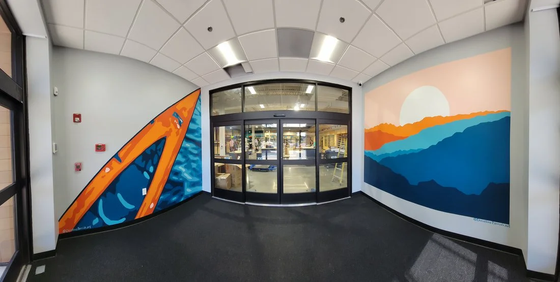 Interior view of a building entrance with large murals on the walls; one mural depicts an orange and blue kayak on water, and the other depicts a stylized sunset over blue mountains.