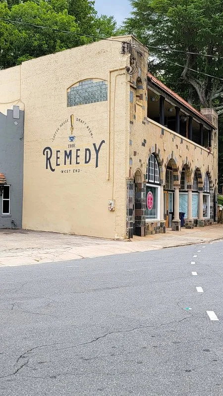 A two-story building with a beige stucco exterior and arched windows, called The Remedy, located on a street corner with trees in the background.