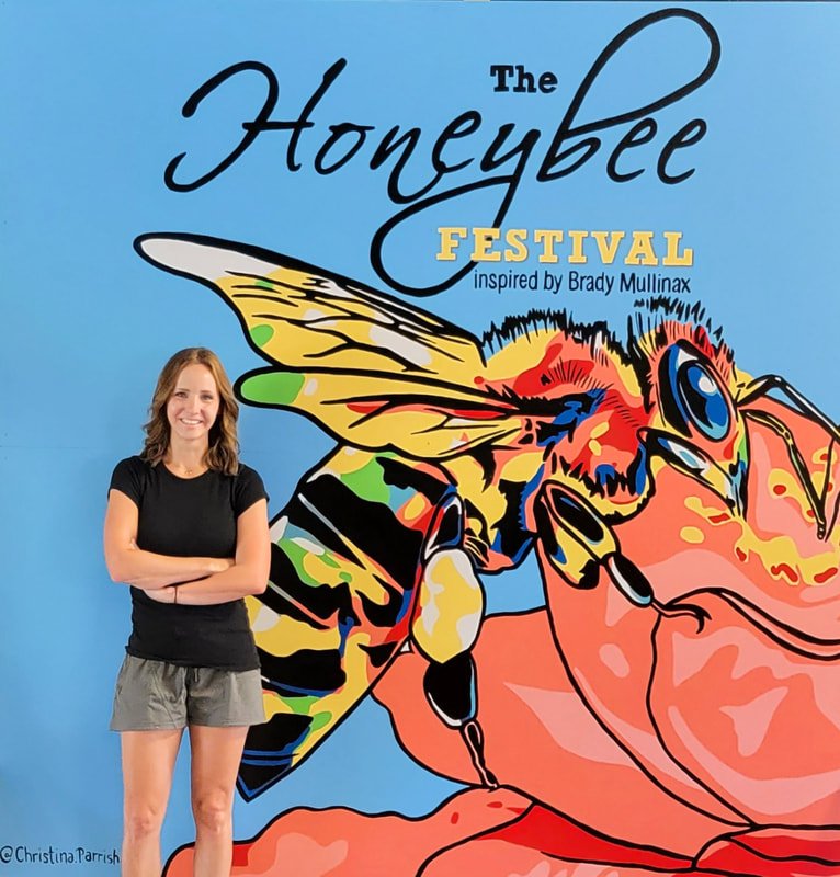 A young woman in a black t-shirt and gray shorts standing with her arms crossed, next to a colorful mural of a bee and a crab, with text about the Honeybee Festival inspired by Brady Mullinax.
