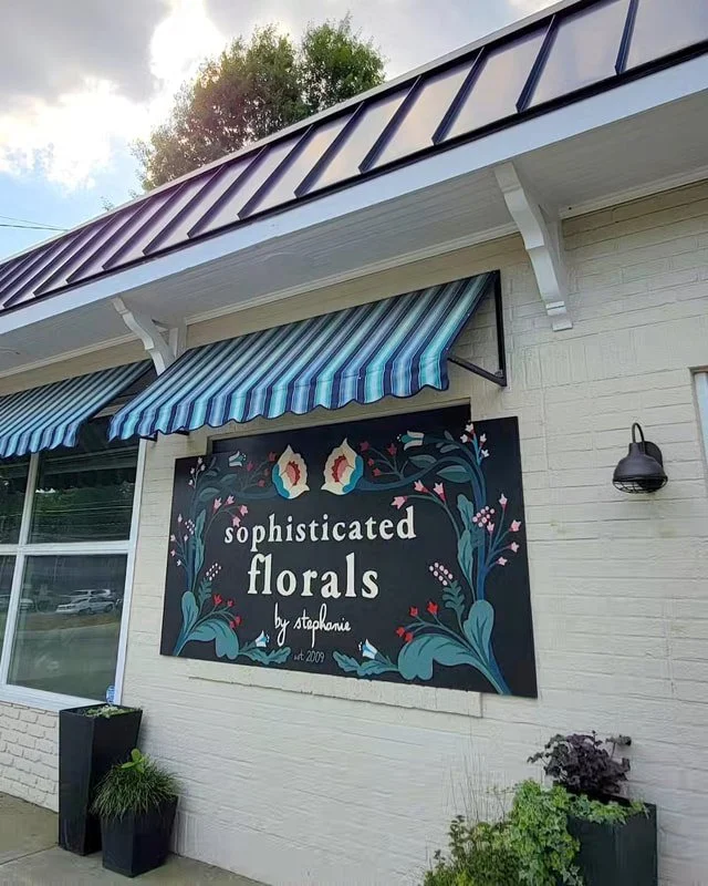 Exterior view of a floral shop called 'sophisticated florals by stephanie' with striped blue awnings and decorative plants in pots outside.