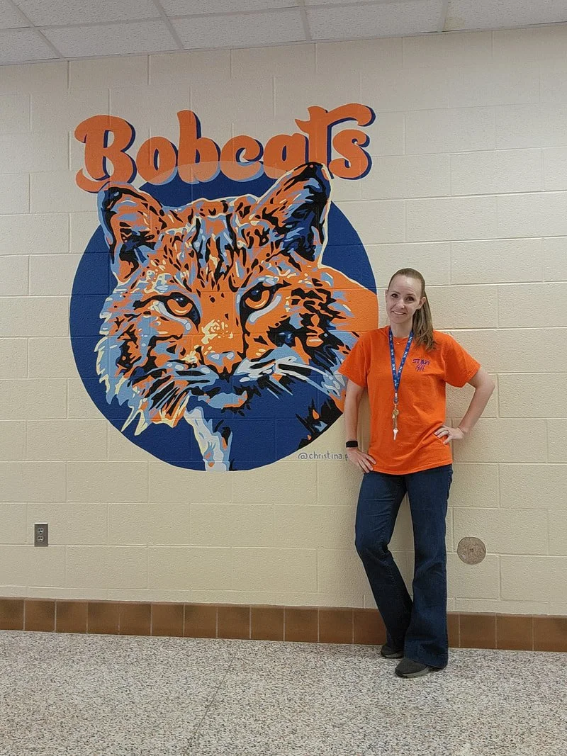 A woman in an orange shirt standing next to a large mural of a bobcat's face on a school wall.
