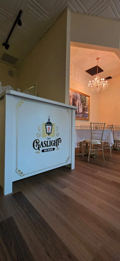 Interior view of a restaurant with a white log-style table or counter, labeled 'The Gaslight on Main,' next to a dining area with white chairs, a chandelier, and a framed picture on the wall.