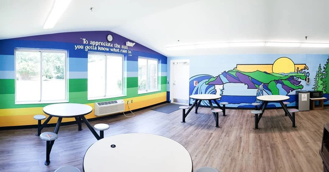 Colorful room with mural of mountains, river, and sun, featuring round tables and squeegee chairs, large windows, and a quote about appreciating sunshine.