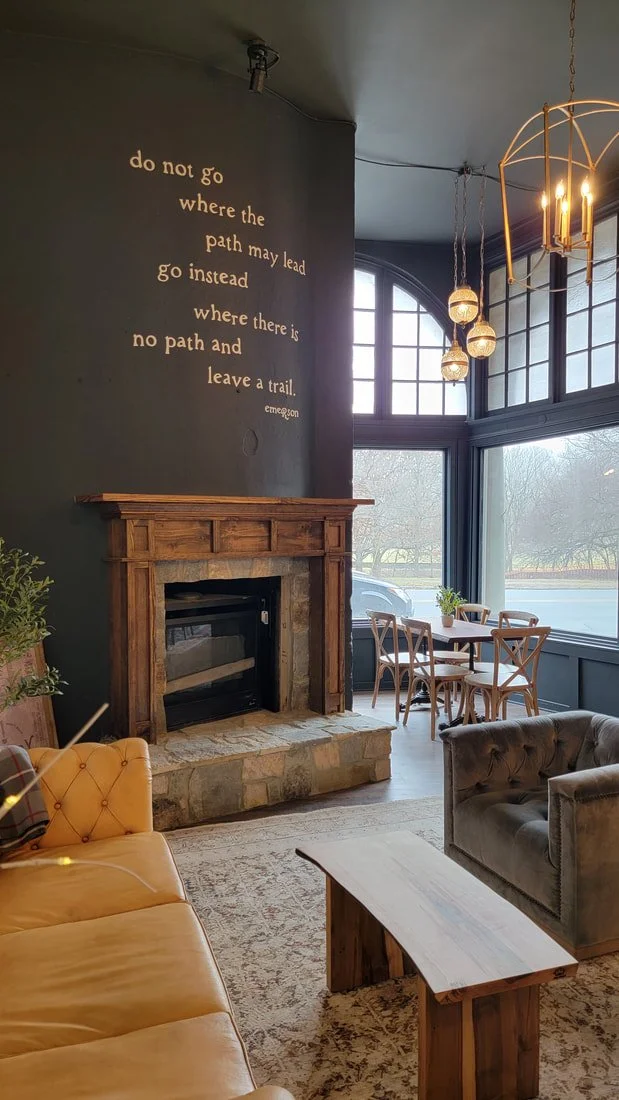 Interior of a cozy living room with a stone fireplace, a yellow tufted sofa, a gray armchair, a wooden table, a round wooden dining table with four chairs, large windows, and a hanging chandelier. A quote on the wall reads: "do not go where the path 