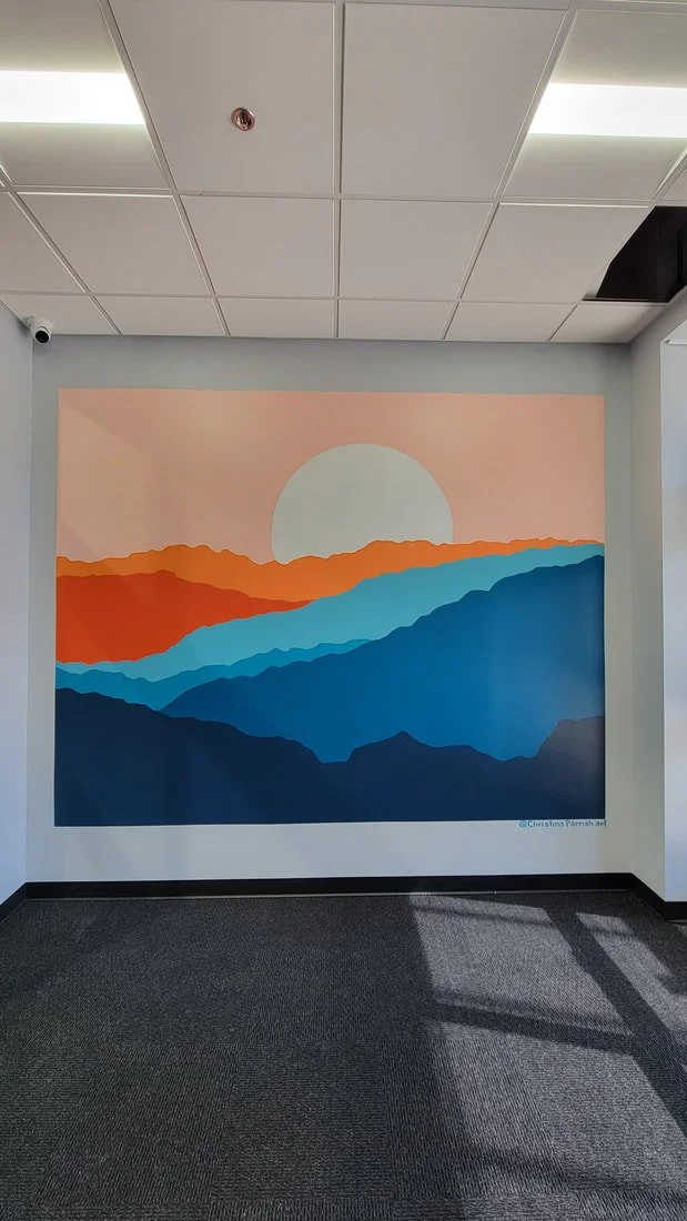 A colorful mural of a sunset over mountain silhouettes in shades of red, orange, blue, and dark blue, on a wall in an indoor space with a drop ceiling.