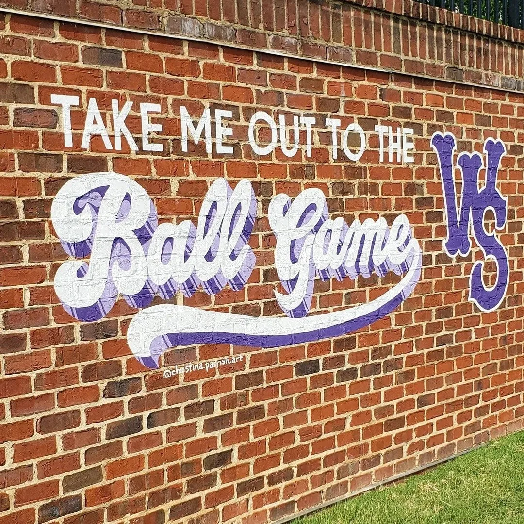 Decorative mural on a brick wall saying, "Take me out to the ball game" with a large purple letter "K".