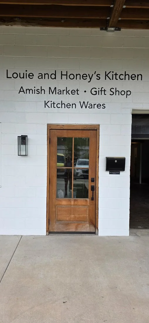 White brick wall with black lettering sign that reads, "Louie and Honey's Kitchen Amish Market · Gift Shop · Kitchen Wares," wooden door with glass window, black mailbox, and sidewalk in front.