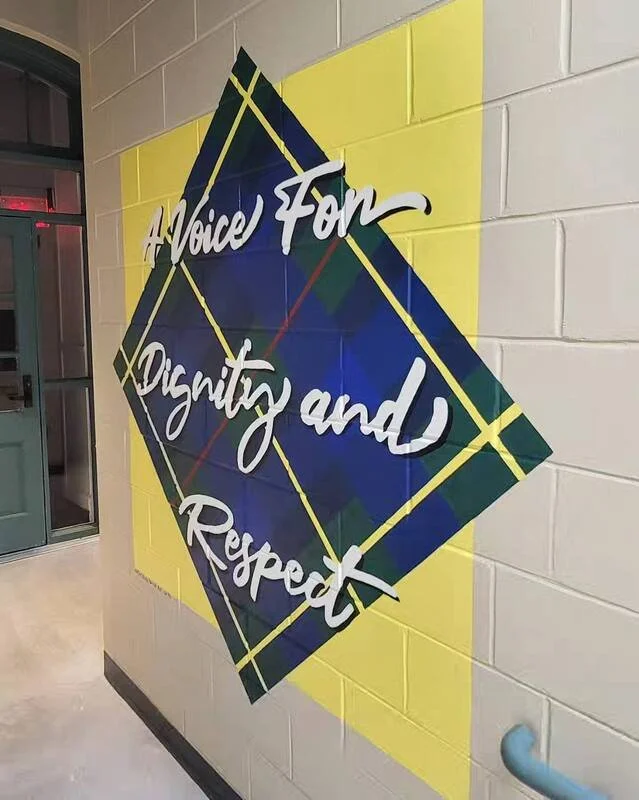 A mural on a white brick wall with the message 'A Voice for Dignity and Respect' written in white cursive text over a dark blue diamond-shaped background with yellow and green accents.
