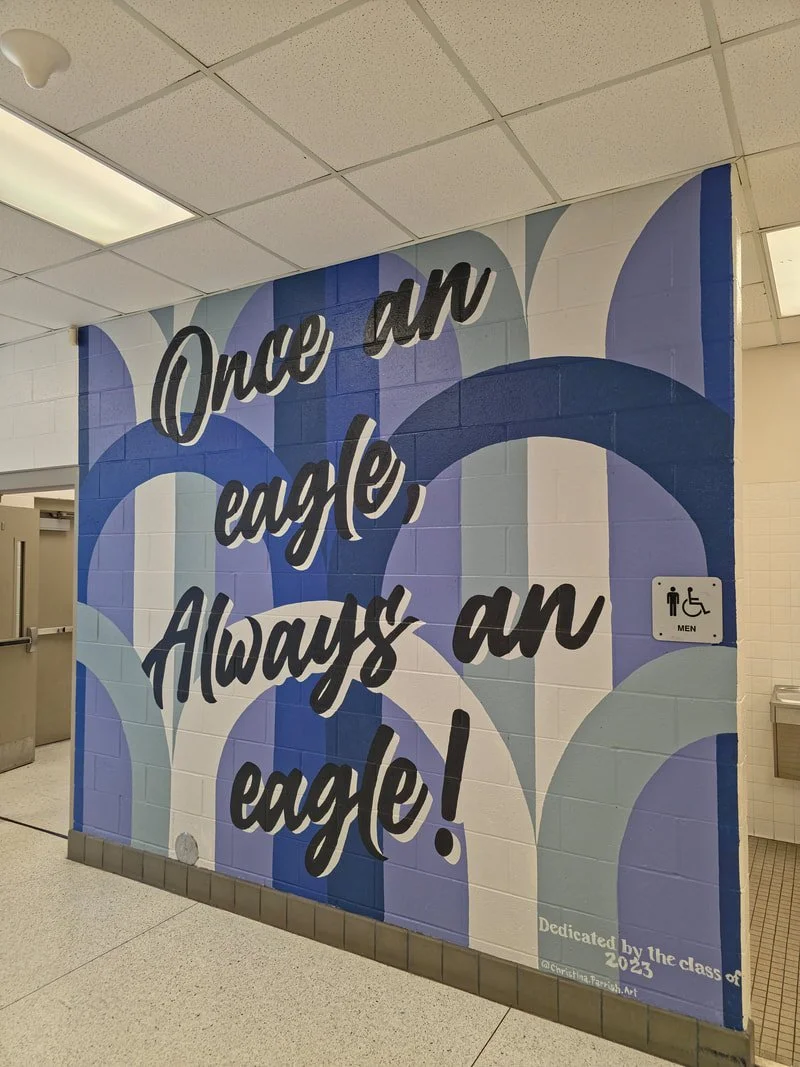 Colorfully painted wall with the quote "Once an eagle, Always an eagle!" in large black script letters. background features abstract blue, purple, and white shapes. Small sign indicates men's restroom nearby.