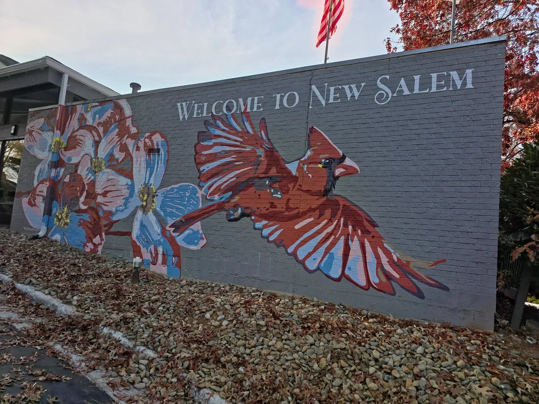 A colorful mural on a brick wall with a bird, flowers, and the text "Welcome to New Salem." An American flag is flying above the mural.