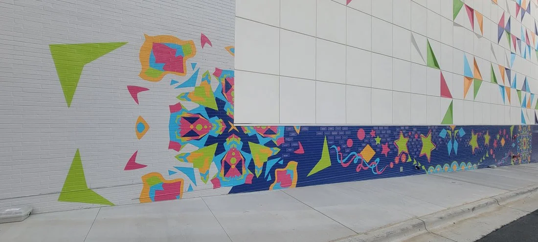 Colorful mural on a white brick wall with various geometric and star-shaped patterns in bright colors, extending along a sidewalk.