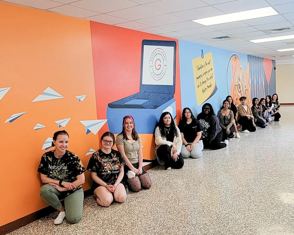 A group of mostly young women sitting on the floor in a line against a colorful wall mural featuring a laptop, paper airplanes, a tiger face, and a quote on a sticky note in a school or community center hallway.