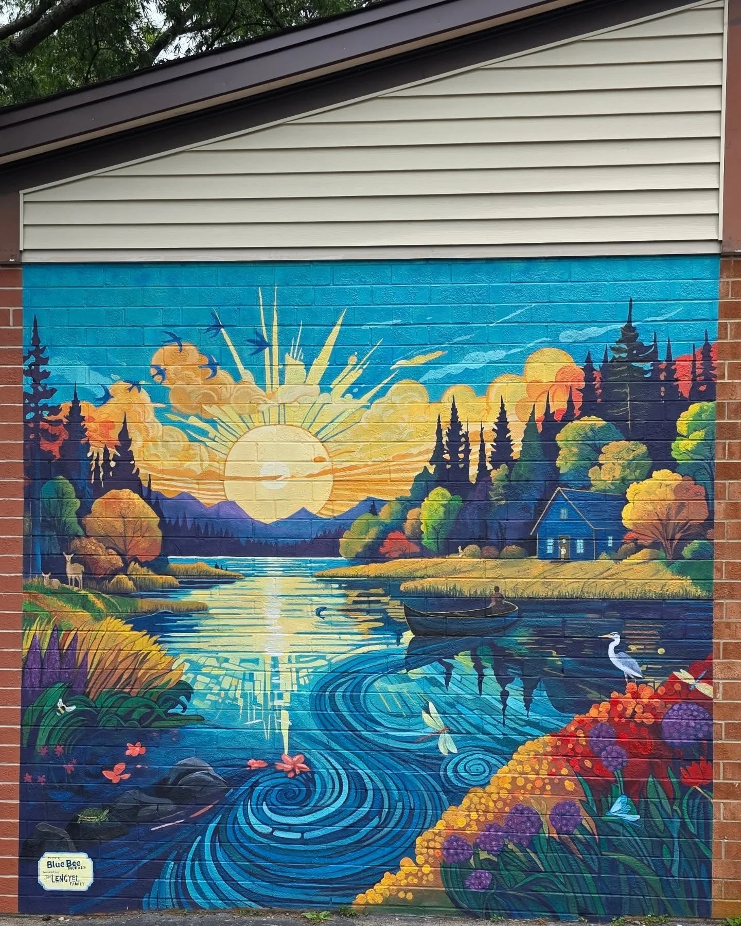Colorful mural depicting a sunrise over a river, with trees, mountains, birds, a person in a boat, and wildlife including a heron, surrounded by colorful foliage.